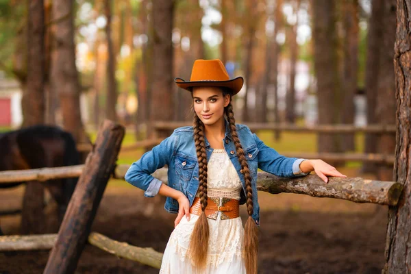 Beautiful cowgirl with extra long braided hair in vintage lace dress and orange hat posing on ranch. Portrait of a pretty model in countryside. Warm art.