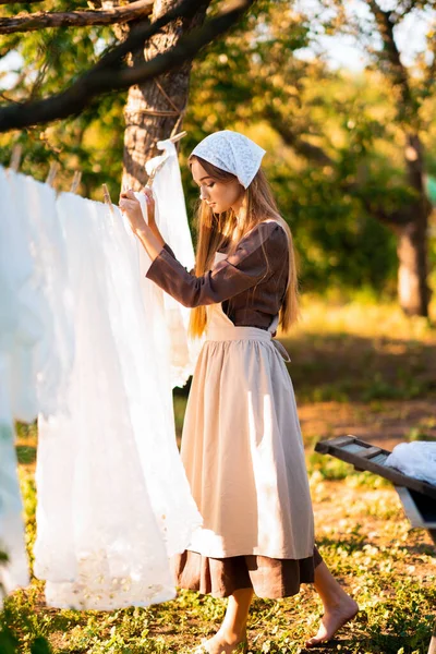 stock image Pretty young laundress in medieval costume washing dresses in old trough on nature in a village.Beautiful girl working in countryside. Fairytale art work.