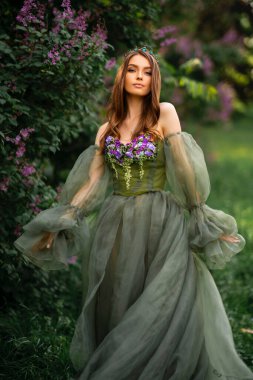 Taht içindeki muhteşem kraliçe ve leylak bahçesinde poz veren güzel yeşil uzun elbiseli. Moda gençliğinin portreleri..