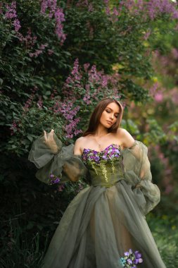 Taht içindeki muhteşem kraliçe ve leylak bahçesinde poz veren güzel yeşil uzun elbiseli. Moda gençliğinin portreleri..