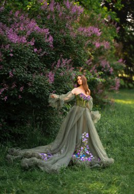 Taht içindeki muhteşem kraliçe ve leylak bahçesinde poz veren güzel yeşil uzun elbiseli. Moda gençliğinin portreleri..