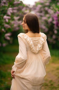 Leylak bahçesinde poz veren uzun saçlı ve klasik beyaz elbiseli güzel bir kız. Romantik model doğanın tadını çıkarıyor..