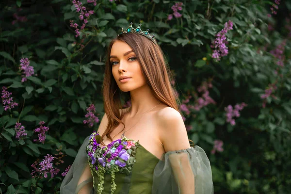 Gorgeous queen in crown and beautiful green long dress with flowers posing in lilac garden. Portraits of fashion young model.