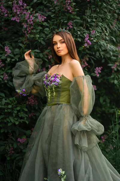华丽的女王头戴皇冠 美丽的绿色长裙 花朵点缀在紫丁香花园里 年轻时尚模特的肖像 — 图库照片