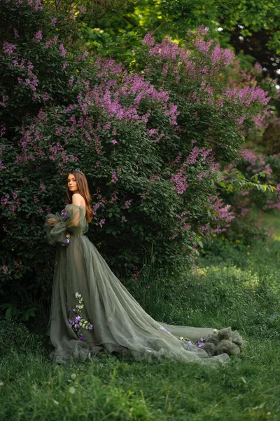 王冠の豪華な女王とライラックガーデンにポーズをとる花と美しい緑の長いドレス ファッション若いモデルの肖像 — ストック写真