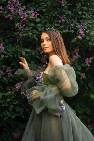 华丽的女王头戴皇冠 美丽的绿色长裙 花朵点缀在紫丁香花园里 年轻时尚模特的肖像 — 图库照片