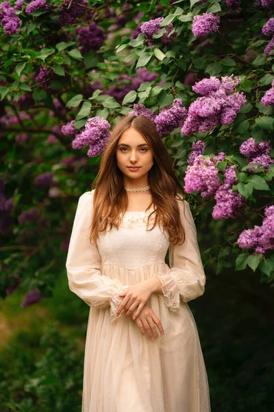 Beautiful girl with long hair and vintage white dress posing in lilac garden. Romantic model enjoying the nature.