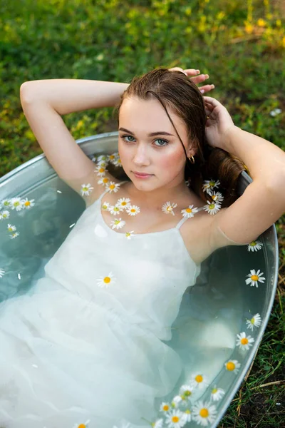 白いドレスの美しいブロンドの長い髪の少女は 水とカモミールで困難に横たわっています 暖かいアートワーク 若いモデルはバックグラウンドとしてヴィンテージの服で自然に風呂に入ります — ストック写真