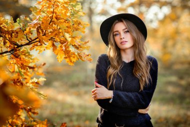 Renkli sonbahar yapraklarının yanında duran koyu renk elbiseli ve siyah şapkalı güzel bir kızın portresi. Romantik bir kadının sanat eseri. Kameraya bakan oldukça hassas bir model..