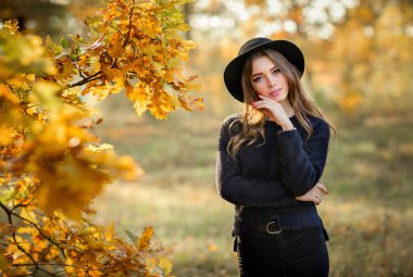 Renkli sonbahar yapraklarının yanında duran koyu renk elbiseli ve siyah şapkalı güzel bir kızın portresi. Romantik bir kadının sanat eseri. Kameraya bakan oldukça hassas bir model..
