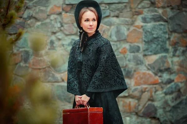 Siyah, eski moda elbise ve kapüşonlu güzel bir kadın taş duvarın yanında duruyor. Valizli romantik bir kadının sanat eseri. Tatlı şefkat modeli rüya görüyor ve uzaklara bakıyor. Seyahat eden kadın.