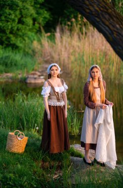 Ortaçağ kostümü giymiş iki çamaşırcı nehir kenarında elbise yıkıyor. Kırsalda çalışan güzel bir kız. Peri masalı eserleri.