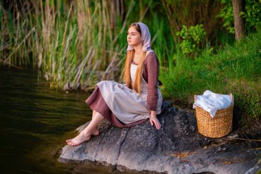 Pretty young laundress in medieval costume standing near river. Fairytale art work. clipart