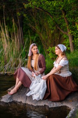 Ortaçağ kostümü giymiş iki çamaşırcı nehir kenarında elbise yıkıyor. Kırsalda çalışan güzel bir kız. Peri masalı eserleri.