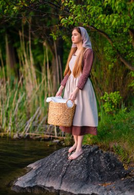 Nehrin yanında duran ortaçağ kostümlü genç çamaşırcı kız. Peri masalı eserleri.