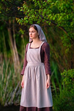 Nehrin yanında duran ortaçağ kostümlü genç çamaşırcı kız. Peri masalı eserleri.