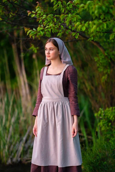 Nehrin yanında duran ortaçağ kostümlü genç çamaşırcı kız. Peri masalı eserleri.