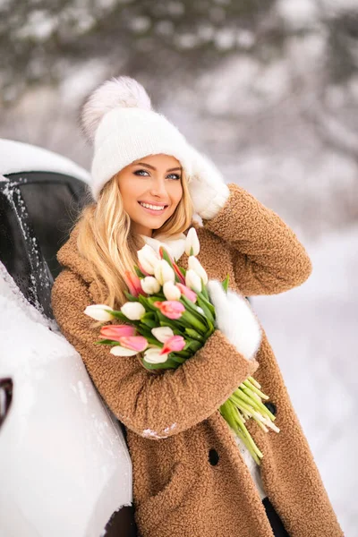 チューリップの花束と暖かい居心地の良い冬の服で美しい女の子は笑って楽しんでいます 白い帽子と明るい冬の日に車の近くにポーズする手袋の若いブロンドモデル スプリングムード — ストック写真