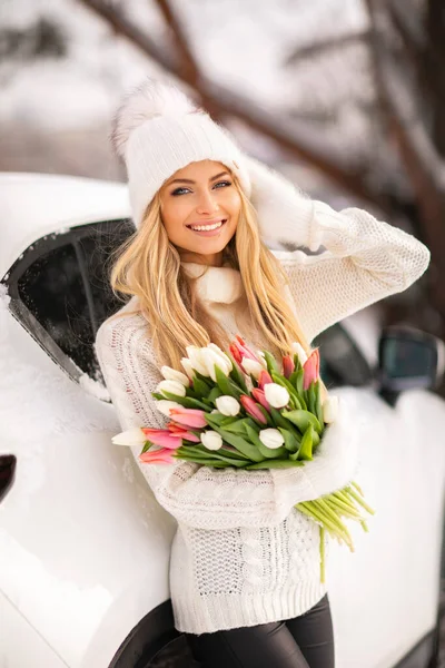 チューリップの花束と暖かい居心地の良い冬の服で美しい女の子は笑って楽しんでいます 白い帽子と明るい冬の日に車の近くにポーズする手袋の若いブロンドモデル スプリングムード — ストック写真