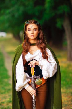 Ortaçağ elf elbisesi içinde güzel kızıl saçlı prenses ve yeşil arka planda savaş pozu veren kılıçlı uzun pelerinli. Diadem 'deki savaşçı hakkında bir masal. Sıcak renkli sanat eserleri.