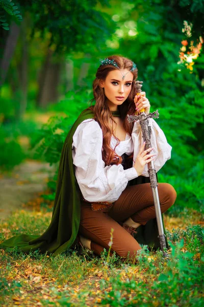 Beautiful red haired princess in medieval elfin dress and long cloak with sword standing in warlike pose on green nature background. Fairy tale story about warrior in diadem . Warm colourful art work.