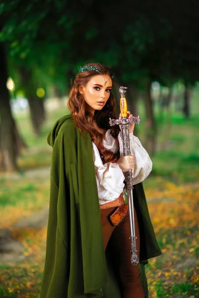 Beautiful red haired princess in medieval elfin dress and long cloak with sword standing in warlike pose on green nature background. Fairy tale story about warrior in diadem . Warm colourful art work.