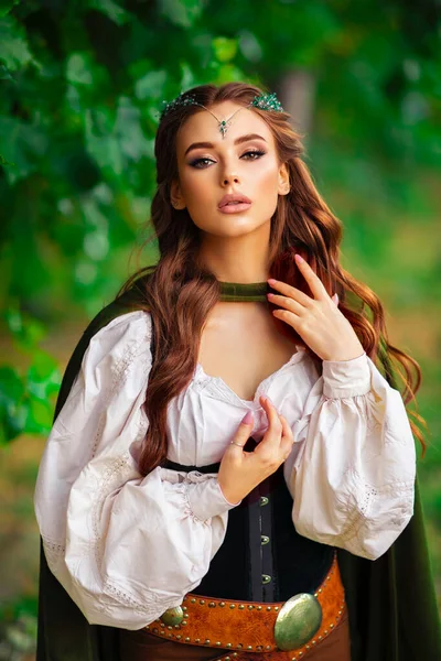 Ortaçağ elf elbisesi ve yeşil arka planda poz veren uzun pelerinli güzel kızıl saçlı prenses. Diadem 'deki savaşçı hakkında bir masal. Sıcak renkli sanat eserleri.