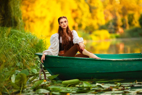美丽的红头发公主穿着中世纪的精灵长裙 坐在一条有绿色背景的河里的小船上 童话故事与温暖多彩的艺术作品 — 图库照片