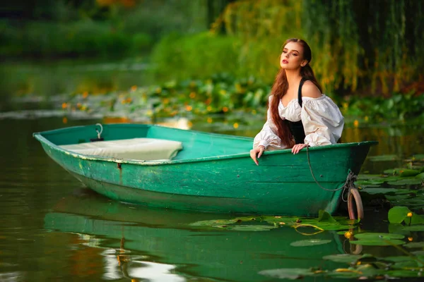 美丽的红头发公主穿着中世纪的精灵长裙 坐在一条有绿色背景的河里的小船上 童话故事与温暖多彩的艺术作品 — 图库照片