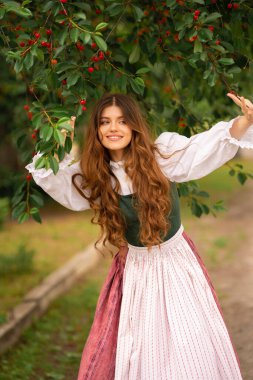 Ortaçağ elbiseli güzel kızıl saçlı kız kiraz ağacının yanında duruyor. Şefkatli, sevimli bir kadınla ilgili peri masalı. Sıcak sanat eserleri..