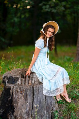 Hasır şapkalı, klasik elbiseli, yeşil arka planda poz veren güzel sarışın kıvırcık kız. Renkli, sıcak sanat eserleri üzerine şapkalı zarif bir bayan.. 