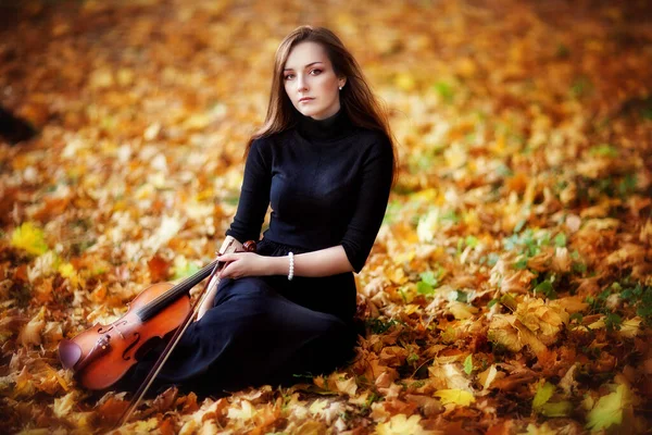 一个穿着深色衣服 带着小提琴的漂亮的黑发女孩的画像 小提琴手坐在秋天的公园里 — 图库照片