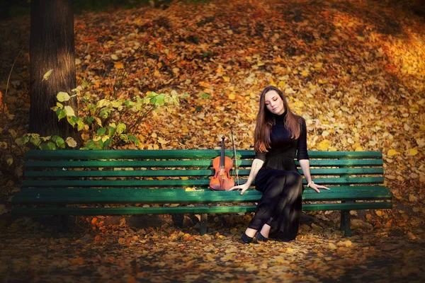 一个穿着深色衣服 带着小提琴的漂亮的黑发女孩的画像 秋天公园里的小提琴家 忧郁地看着黄色的背景 — 图库照片