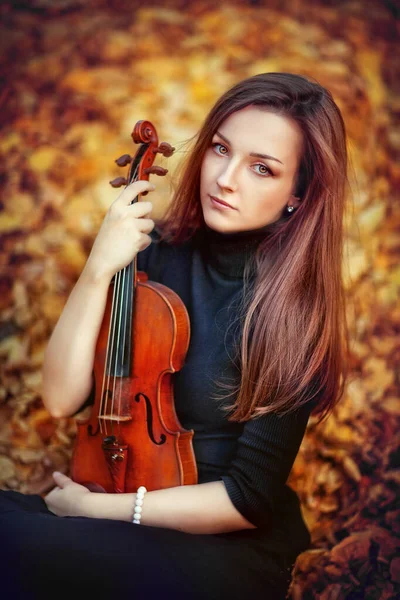 一个穿着深色衣服 带着小提琴的漂亮的黑发女孩的画像 在黄色背景上有着忧郁眼睛的小提琴家 年轻模特儿看着秋天公园里的相机 — 图库照片
