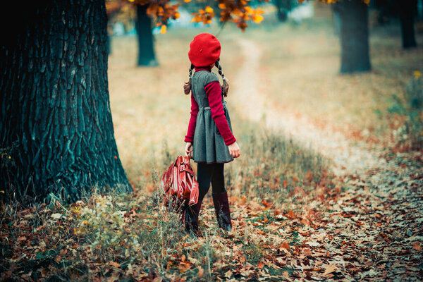 Autumn portrait of a beautiful young schoolgirl in red beret and braid hairs with school bag . Tenderness positive child with bright smile enjoying nature in park.