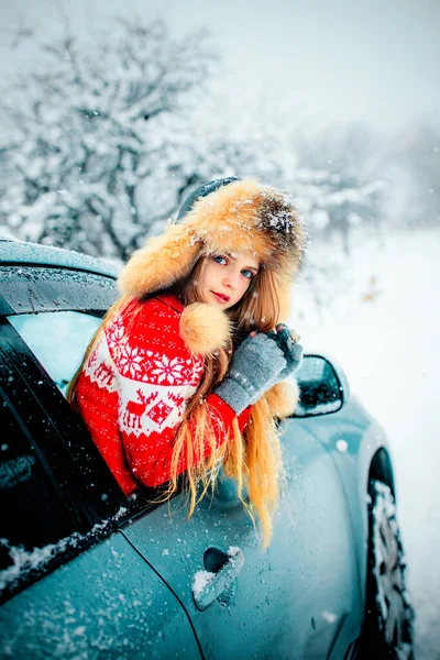 冬雪中美丽的金发姑娘 蓝眼睛 身穿红色毛衣 头戴毛皮帽 站在车旁 — 图库照片