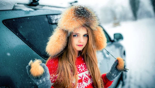 冬雪中美丽的金发姑娘 蓝眼睛 身穿红色毛衣 头戴毛皮帽 站在车旁 — 图库照片