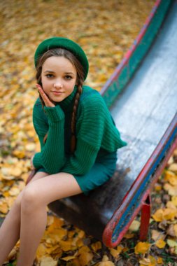 Yeşil bereli ve örgülü güzel bir kız oyun parkında sonbahar doğasının tadını çıkarıyor. Sarı arka planda yapraklarla poz veren sevimli bir genç..