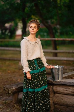 Kırsal kesimdeki uzun elbiseli güzel sarışın kız ellerinde kova dolusu suyla bir köyde poz veriyor. Kırsal kesimdeki sanat fotoğraflarında oldukça genç bir model.. 