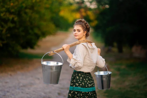 Beautiful  blonde braided girl in rural long dress posing in a village with buckets of water. Pretty young model in countryside art photo.