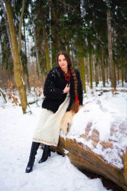 Ukrayna geleneksel elbisesi, siyah kürk mantosu ve kış ormanlarında poz veren kırmızı boncuklu ekstra uzun örülü saçları olan güzel bir kız. Kış arka planında çekici bir kadının portresi..