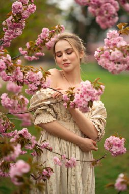 Bahar parkında pembe çiçek çiçeklerinin yanında eski dantel elbiseli güzel sarışın kız. Şefkatli romantik model.