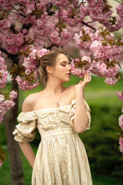 Bahar parkında pembe çiçek çiçeklerinin yanında eski dantel elbiseli güzel sarışın kız. Şefkatli romantik model.
