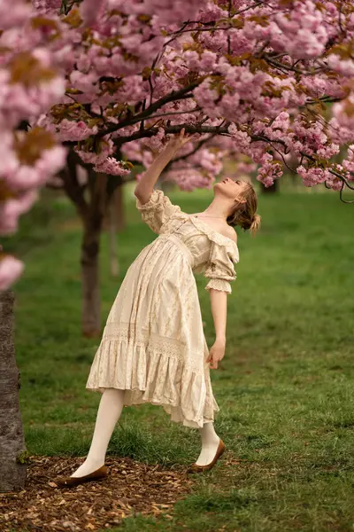 漂亮的金发姑娘穿着老式花边裙站在春天公园的粉红花朵旁 温柔浪漫的造型 图库图片