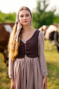 Beautiful blonde girl in scarf and  medieval rural clothes on a pasture with a cows. Farm life.Young model posing on a ranch.Warm art work. clipart