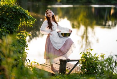 Beautiful red hair girl in white rural dress posing near river with laundry. Warm art work. Young laundress on green background. clipart