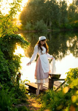 Beautiful red hair girl in white rural dress posing near river with laundry. Warm art work. Young laundress on green background. clipart