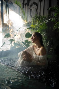Güzel sarışın, uzun saçlı, beyaz elbiseli yeşil bitkilerle gölde poz veren kız. Ophelia hakkındaki peri masalı. Sıcak sanat eserleri.