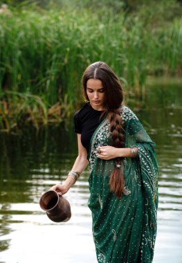 Nehrin kenarında sürahiyle poz veren, ekstra uzun örgülü, Hint geleneksel elbiseli güzel esmer kız. Yeşil doğa arka planında, sari içinde genç ve çekici bir kadının portresi..