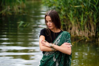 Nehir kenarında poz veren, ekstra uzun örgülü, Hint geleneksel elbiseli güzel esmer kız. Yeşil doğa arka planında, sari içinde genç ve çekici bir kadının portresi..